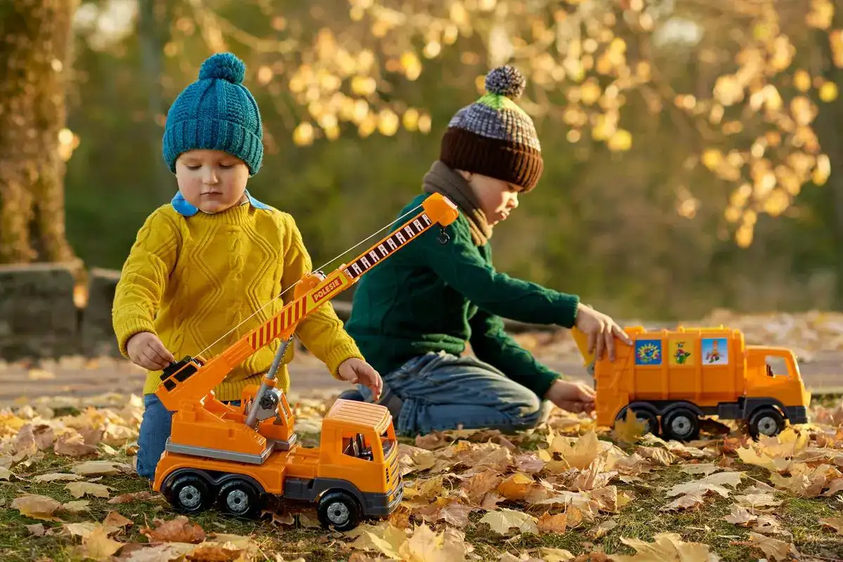 Two young children in warm clothing play with toy construction trucks outside on fallen autumn leaves.