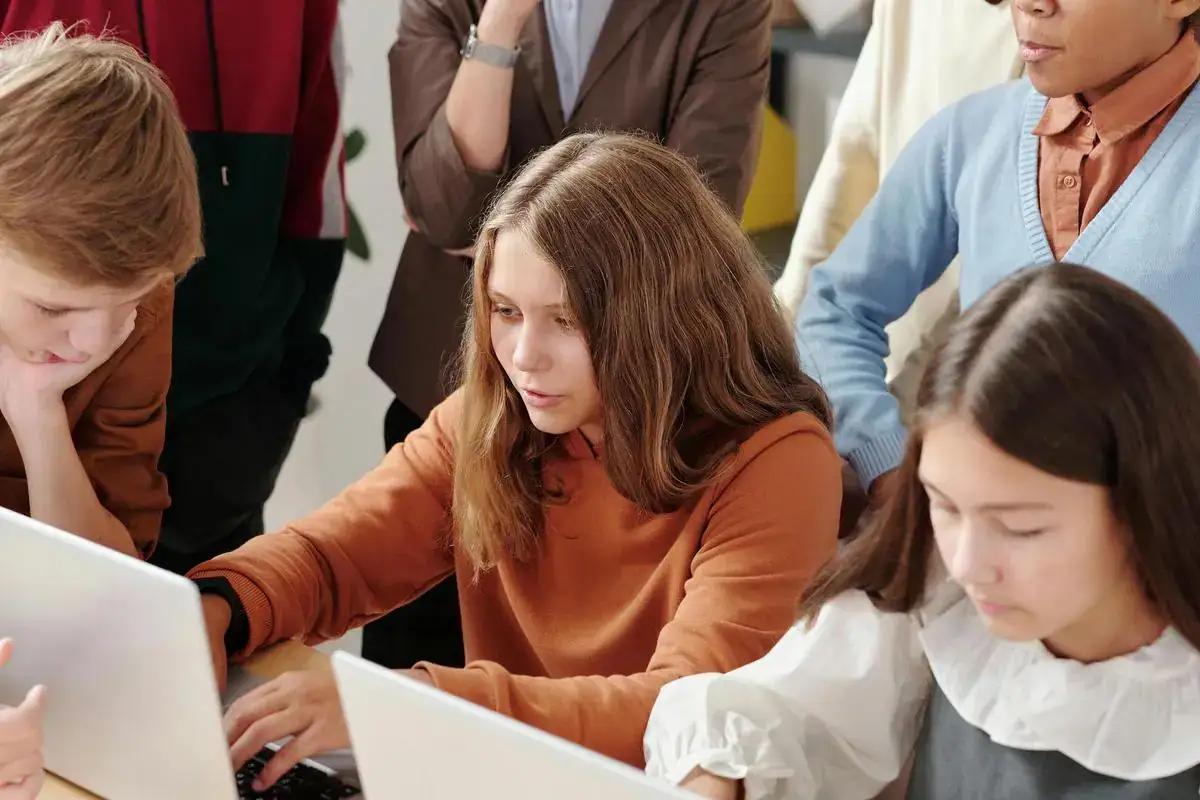 Several students are gathered around laptops, focused on their work in a classroom or group setting.